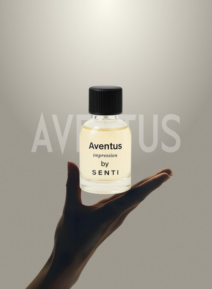 A hand delicately holding a clear glass bottle of Senti Aventus Impression perfume, with the word AVENTUS subtly visible in the background. Highlighting the signature fragrance.
