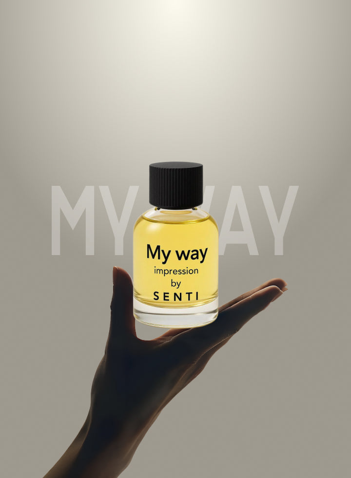 A hand gently presenting a clear Senti 'My Way' Impression perfume bottle, with the text 'MY WAY' partially visible in the background. Second hero image for the fragrance.
