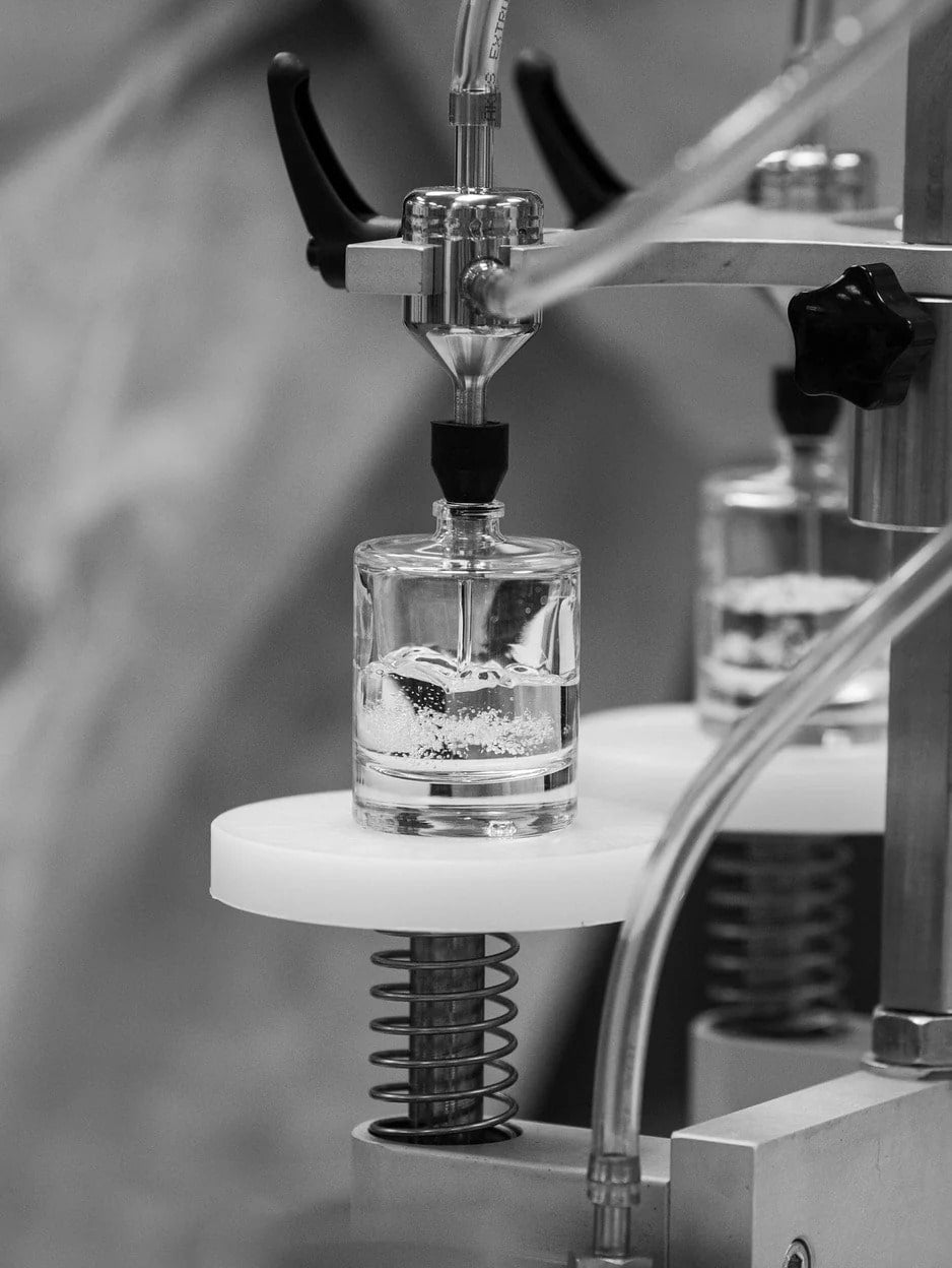 Black and white close-up of a Senti perfume bottle being filled by machinery, showing the precision and quality of the fragrance making process.